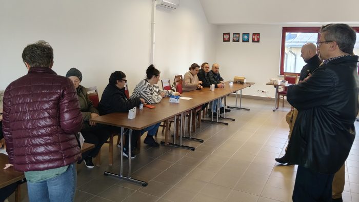 Mieszkańcy gminy uczestniczą w spotkaniu dotyczącym bezpieczeństwa pożarowego. Siedzą przy stole, na którym leżą opakowania z czujkami dymu. Spotkanie odbywa się w sali konferencyjnej z widocznymi symbolami samorządowymi na ścianie.