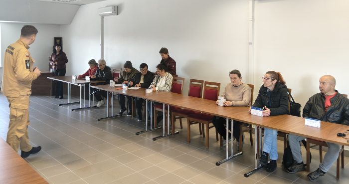 Szkolenie dla mieszkańców z zakresu bezpieczeństwa pożarowego. Strażak tłumaczy zasady działania czujek tlenku węgla i dymu grupie osób siedzących przy stole w sali gminnej